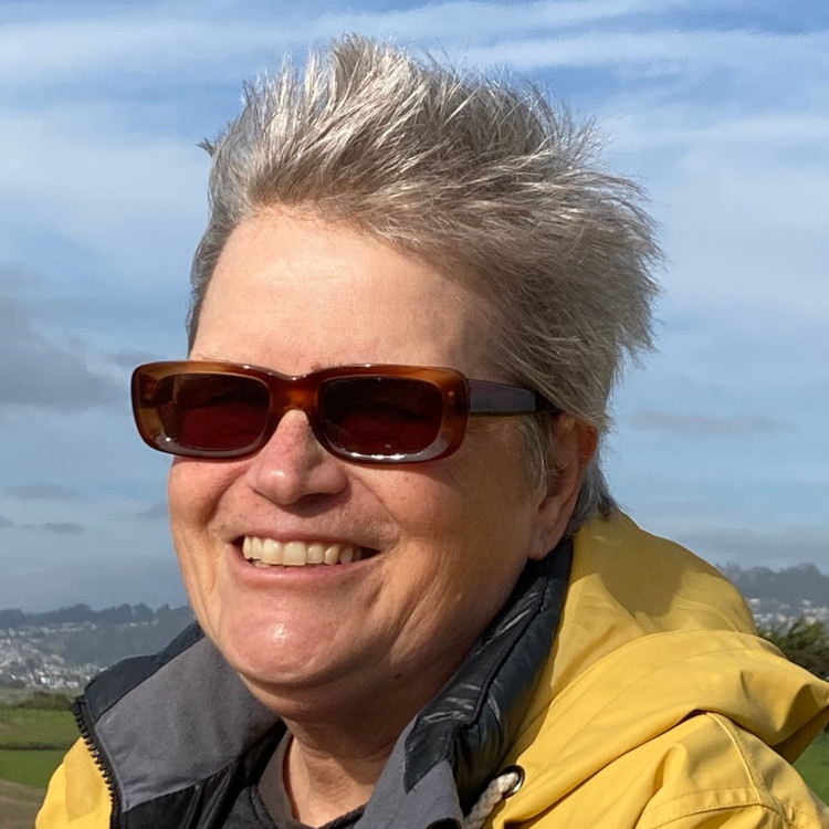 Outdoor portrait of smiling white woman with short windswept silver-blond hair wearing sunglasses and a yellow rain slicker.