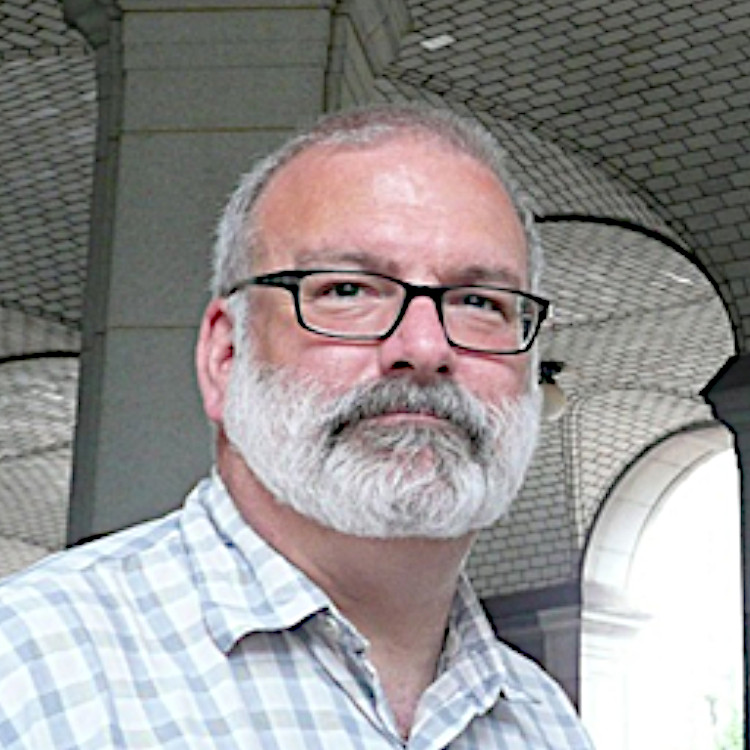 Interior portrait of an older white man with silver-white short hair and a beard wearing an open-collar shirt and glasses.