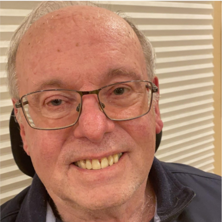 Indoor photo of smiling older white man with glasses and grey hair with dark shirt and fleece vest